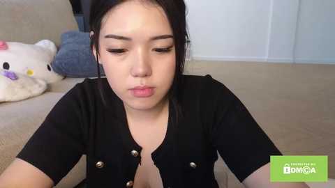 Video of a young Asian woman with straight black hair, wearing a black blouse, blowing a kiss in a beige room with plush toys and white walls.