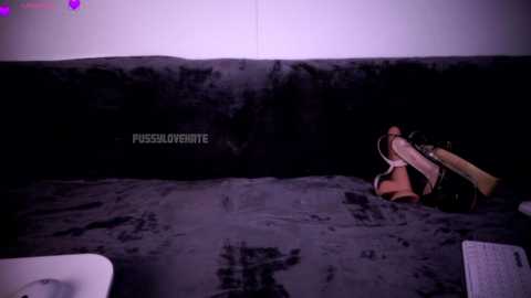 Video of a woman in a black latex bodysuit, lying on a dark purple bed with a black wall behind. She has brown hair and is holding a large, realistic-looking dildo. Text \"PUSAMORENTE\" is visible.