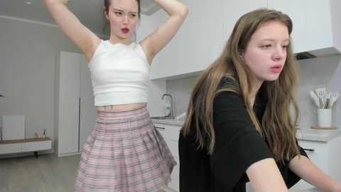 Video of a young woman with fair skin and brown hair in a modern kitchen, wearing a white crop top and pink plaid skirt, looking surprised.