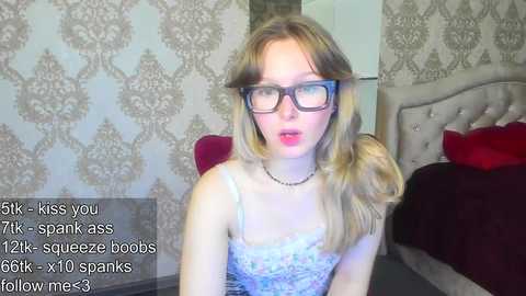 A video of a fair-skinned, blonde woman with glasses, wearing a floral lace bra and a black necklace, in a bedroom with gray damask wallpaper, a bed with red pillows, and text overlay.