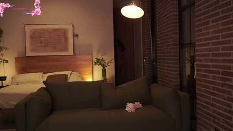 Video of a dimly lit, modern bedroom with a brick wall, wooden headboard, green sofa, white bed, framed artwork, and a pink flower on the floor.