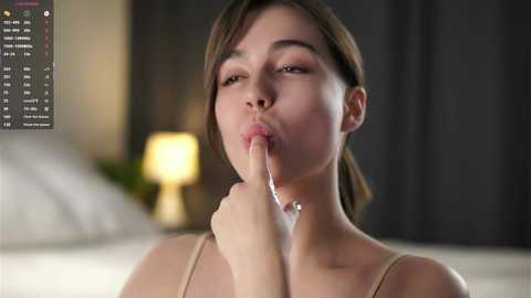 Video of a young woman with fair skin and brown hair, seductively licking her index finger, wearing a beige bra, in a dimly lit bedroom with blurred background.