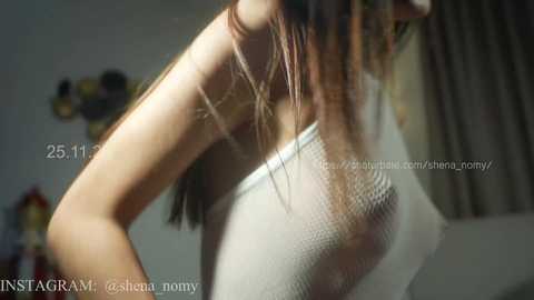 A video of a woman with long brown hair, wearing a sheer white mesh top, leaning forward, showing her back and side. The background is blurry, possibly indoors.