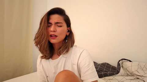 Video of a young woman with shoulder-length brown hair, wearing a white shirt, lying on a bed with a beige blanket and plaid pillow, looking distressed.