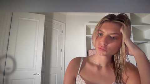 A video of a young, slim, light-skinned woman with wet, blonde hair, wearing a white lace bra, standing in a brightly lit room with white walls and closed doors.