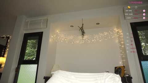 A cozy bedroom with white walls, a white bed, and string lights. Black-framed windows on either side.