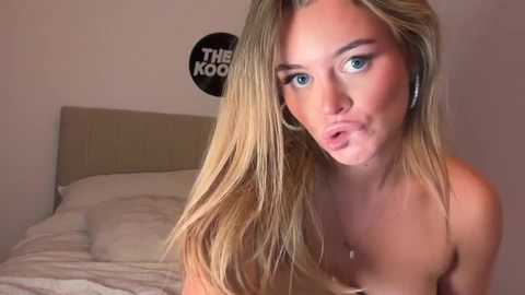 Video of a topless, fair-skinned, blonde woman with blue eyes, puckering her lips in a bedroom with beige walls and a bed.