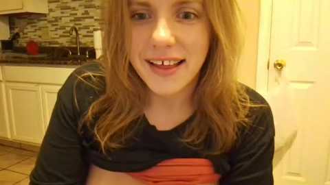 Video of a young woman with light skin, shoulder-length brown hair, and a friendly smile. She's wearing a black top and a red shirt, in a modern kitchen with white cabinets, a mosaic tile backsplash, and a closed door.