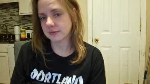 Video of a young Caucasian woman with light skin and shoulder-length brown hair, wearing a black \"DORTLEMAN\" shirt, standing in a modern kitchen with white cabinets, a backsplash, and a white door.