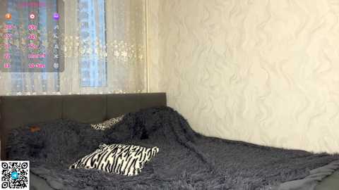 Video of a modern bedroom with a dark gray, textured bedspread, a zebra-patterned pillow, and a light cream wallpaper with a subtle, wavy design. A large window with sheer curtains displays colorful city lights.