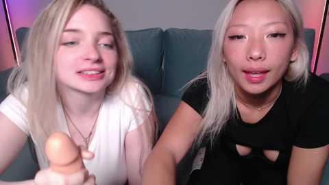 Video of two young, fair-skinned women with long blonde hair, wearing white and black tops, holding a realistic sex doll.