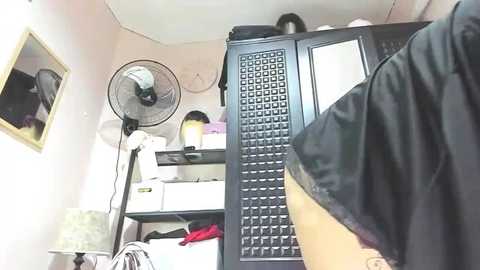 A video of a cluttered bedroom with a white wall, black clothes rack, black fan, white shelves, and a black and white striped shirt hanging in the foreground.