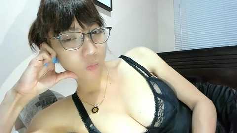 Video of an Asian woman with short, dark hair, glasses, and a black lace bra, sitting on a bed. She has a light skin tone and a slender physique. The room has white walls, a window with closed blinds, and a dark wooden headboard.