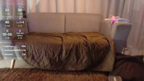 Video of a beige couch with a brown, textured blanket, positioned against light-colored curtains. A blue chair with a pink cushion sits to the right.