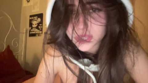 Media: Video of a topless young woman with long, dark hair, wearing a white hood, in a bedroom with a metal bed frame, a poster, and a plush toy.