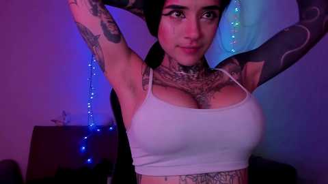 Video of a tattooed woman with medium skin tone, dark hair, and large breasts, wearing a white crop top, posing with arms raised against a purple-lit background.