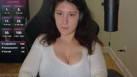 A video of a fair-skinned, dark-haired woman with large breasts, wearing a white top, sitting in a black gaming chair, with a live-streaming overlay showing viewers and chat.
