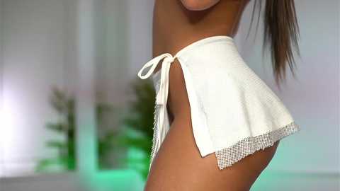 Video of a fit, tan-skinned woman in a white, open-back, pleated skirt, with a blurred background featuring green plants and soft lighting.