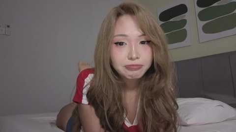 Video of an Asian woman with long, wavy blonde hair, light skin, and a slim physique, wearing a red top. She lies on a white bed in a minimalist bedroom with green abstract art on the wall.