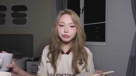 A video of a young Asian woman with long, wavy blonde hair and pale skin, wearing a white soccer jersey, eating noodles in a dimly lit bedroom.