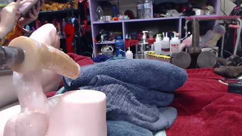 Media: Video of a messy bedroom with a pink dildo being used, surrounded by various sex toys and toiletries on a blue bedspread.