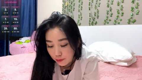 A video of an Asian woman with long black hair, wearing a white blouse, lying on a pink bed, surrounded by green ivy wall decor and blue curtains.