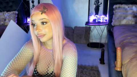 Video of a young woman with long, straight, pastel pink hair and light skin, wearing a fishnet top, in a dimly-lit, cozy room with purple neon signs and a large dildo.