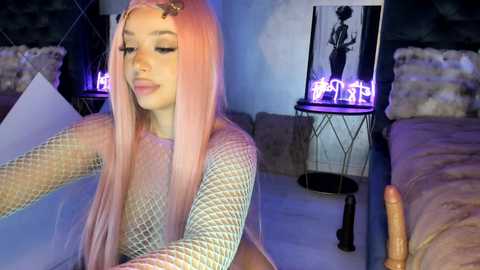Video of a young woman with long, pastel pink hair, wearing a fishnet top, posing seductively on a bed with a furry pillow, surrounded by neon lights.