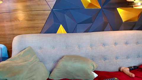 Video of a modern, geometric-patterned wall with dark blue and yellow triangular shapes behind a light gray, tufted headboard with a plush green pillow and a red blanket.