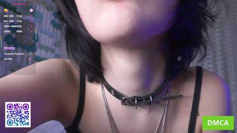 A video of a young woman with pale skin, black hair, and a black leather choker with a metal chain, wearing a black tank top. The background features a blurred, colorful mural.