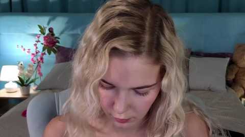 Video of a blonde woman with wavy hair, wearing a sleeveless top, sitting in a dimly lit bedroom. Background includes a bed with pillows, a nightstand with a lamp and flowers, and a blue wall with a floral pattern.