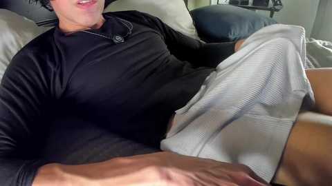 Video of a light-skinned man with short dark hair, wearing a black long-sleeve shirt and grey shorts, reclining on a grey couch, with a visible erection.