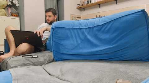 Video of a bearded man in shorts, barefoot, typing on a laptop on a blue and gray sectional sofa in a cozy living room with wooden shelves and a floral arrangement.