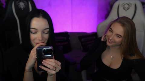 Video of two young women in a dimly lit room, one taking a selfie with a phone, the other smiling in the background.
