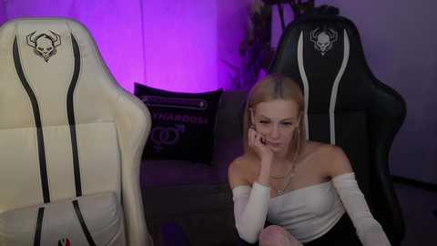 Video of a blonde woman with glasses, wearing a white off-shoulder top and black skirt, sitting in a black gaming chair, indoors with purple lighting and gaming gear in the background.