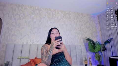 Video of a woman with long black hair, tattoos, wearing a green striped dress, taking a selfie in a modern bedroom with a white headboard, green plants, and a colorful background.