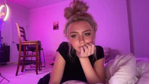Video of a blonde woman with a high bun, wearing a black top, resting on a bed with a pinkish glow, in a cozy, dimly lit room.
