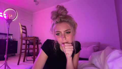 Video of a young woman with light skin and blonde hair in a messy bun, wearing a black top, kneeling on a bed. She is blowing into her hand. Room lit by pink and purple LED lights, with a wooden chair and table in the background.