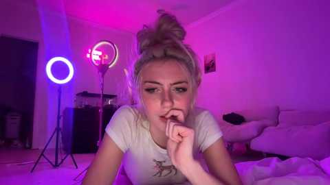 A video of a young woman with light skin, platinum blonde hair in a messy bun, wearing a white t-shirt and biting her finger, lying on a bed. The room is lit with pink and purple LED lights, and there's a ring light and a speaker in the background.