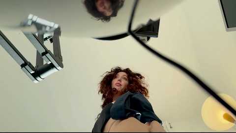 A video of a woman with curly brown hair, wearing a black top, standing in a modern, minimalist room with a glass ceiling and light fixtures.