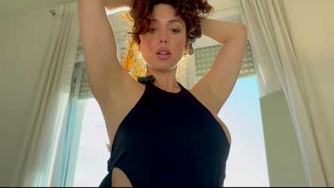 Video of a curly-haired woman with light skin, wearing a black halter top, standing in a bright room with white curtains.