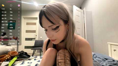 Video of a young Asian woman with long, straight hair and dark eye makeup, seated on a bed, wearing a black choker. Background features a messy room with a closed door, chair, and scattered items.