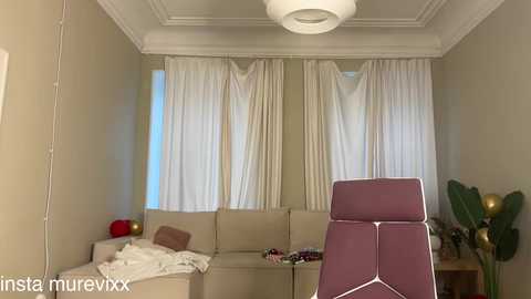 Video of a modern living room with beige walls, white curtains, a beige sectional sofa, a pink recliner chair, a green potted plant, and a round white ceiling light.