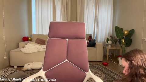 Video of a woman in a beige living room with a pink, geometric recliner chair, beige couch, white curtains, and potted plants.