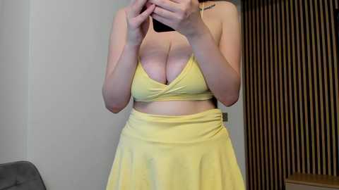 Video of a fair-skinned woman with medium-sized breasts, wearing a yellow, low-cut, halterneck top and matching skirt. She holds a phone in front of her face, partially obscuring it. Background includes a grey couch and a wooden-paneled wall.
