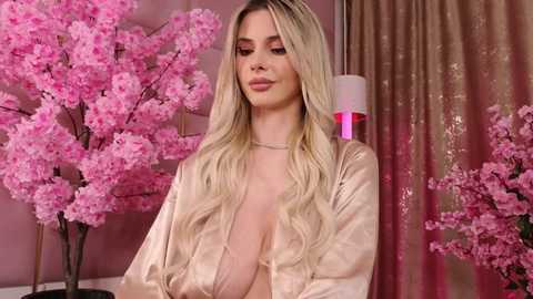 Video of a blonde woman with long, wavy hair, wearing a satin, light pink robe, standing in a room with pink cherry blossom trees and gold curtains, giving a serene, elegant vibe.