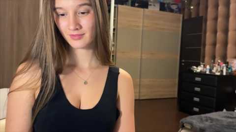 Video of a fair-skinned, long-haired young woman in a black tank top, smiling slightly, in a cluttered, dimly lit room with wooden furniture, shelves, and a bed visible in the background.