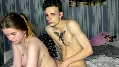 Video of a nude young woman with light skin and long brown hair, sitting on a bed, and a nude young man with a tattoo on his chest, standing behind her.
