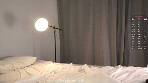 A video of a minimalist bedroom featuring a neatly made bed with white sheets, a single bulb lamp, and a large, gray curtain. A digital clock on the wall displays the time.