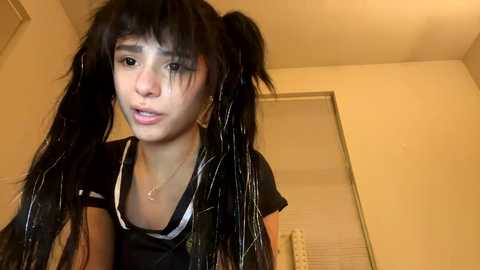 Video of a young woman with long black pigtails, wearing a black top, standing in a dimly lit room with beige walls and a window.